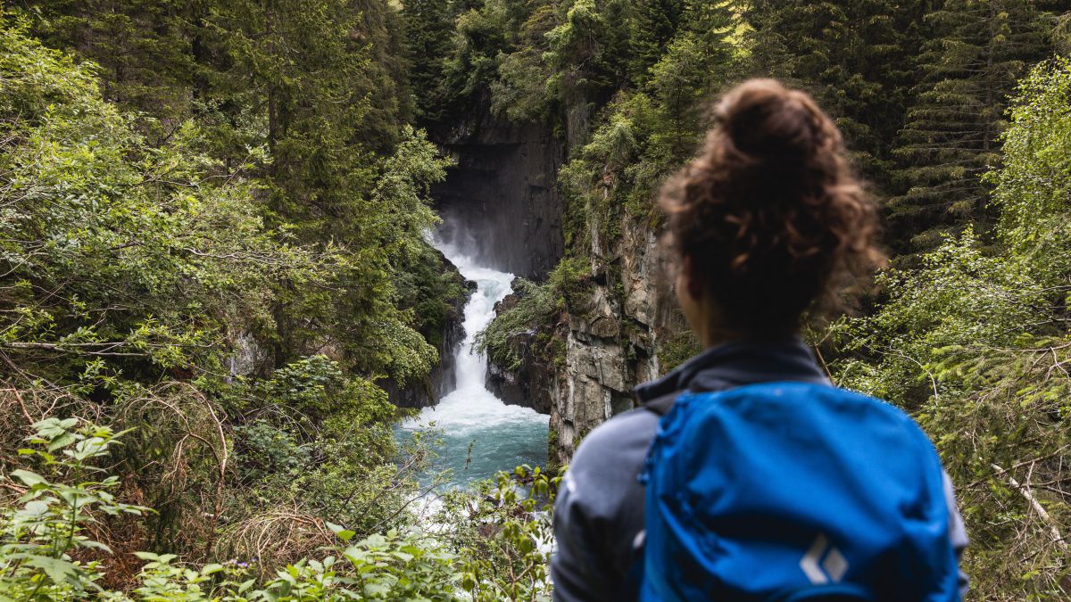 Wir stehen hinter einer Frau mit Wanderrucksack, sie blickt auf einen Wasserfall, der schäumend weiß in den blaugrünen Fluß fällt. Rundherum grün-braune Waldwildnis und felsiges Ufer.