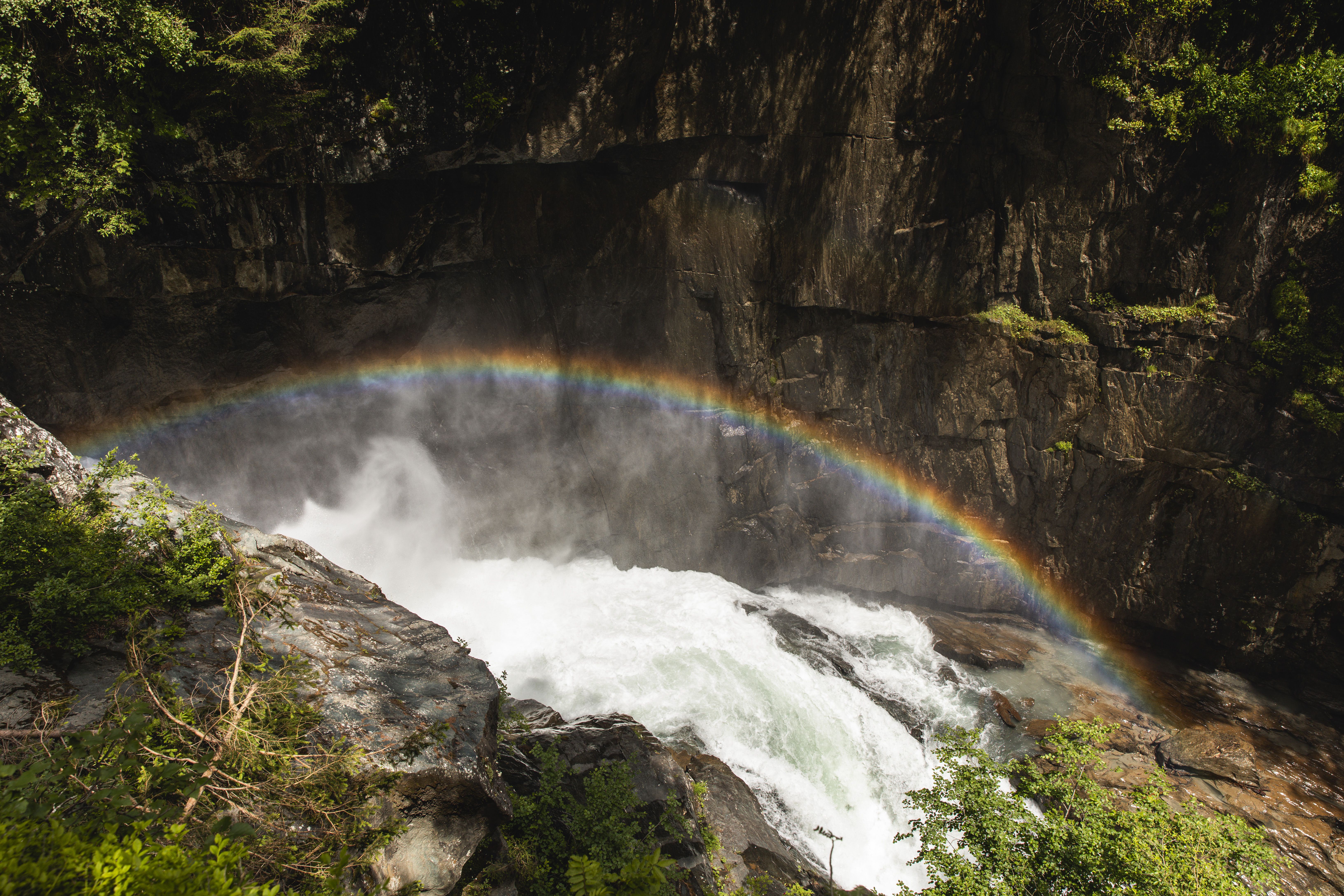 In die Gischt über dem tosenden Wasser des Flusses malt das Sonnenlicht einen bunten Regenbogen an die graue Felswand.