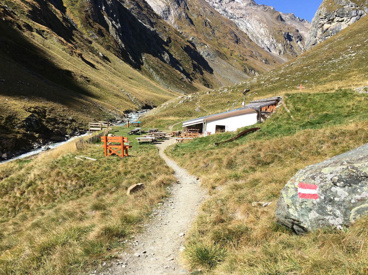 Ein steiniger Pfad führt bergab, am Wegesrand eine rot-weiß-rote Markierung, vor uns drückt sich eine Berghütte mit flachem Dach an den Wiesenhang. Links unten verläuft der Fluss. Auf beiden Seiten geht es steil in die Höhe, wir sind inmitten der Berge.