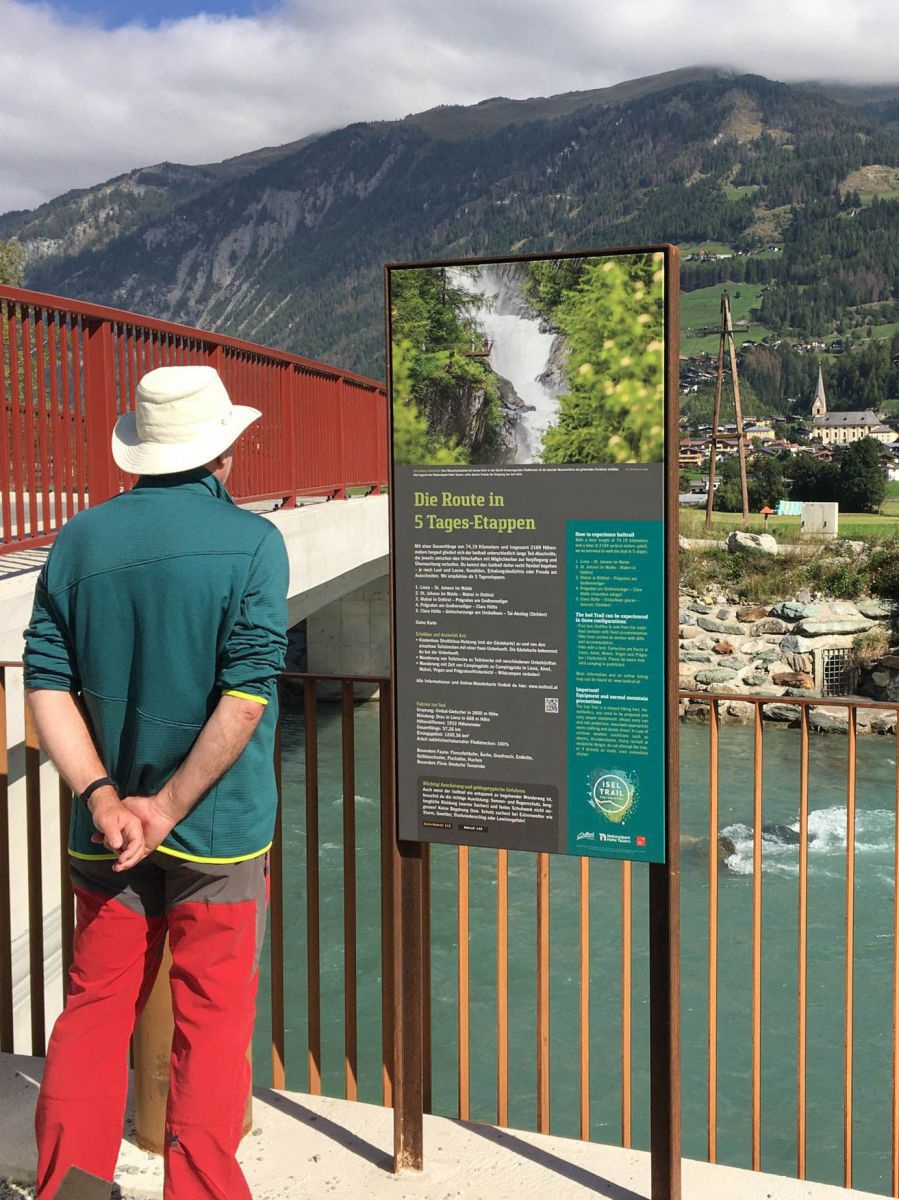 Ein Mann mit Sonnenhut steht auf einer Brücke vor einer Infotafel und liest, die Hände am Rücken verschränkt. Hinter dem roten Geländer sieht man weiter unten den Fluss. Am Horizont bewaldete Berghänge.