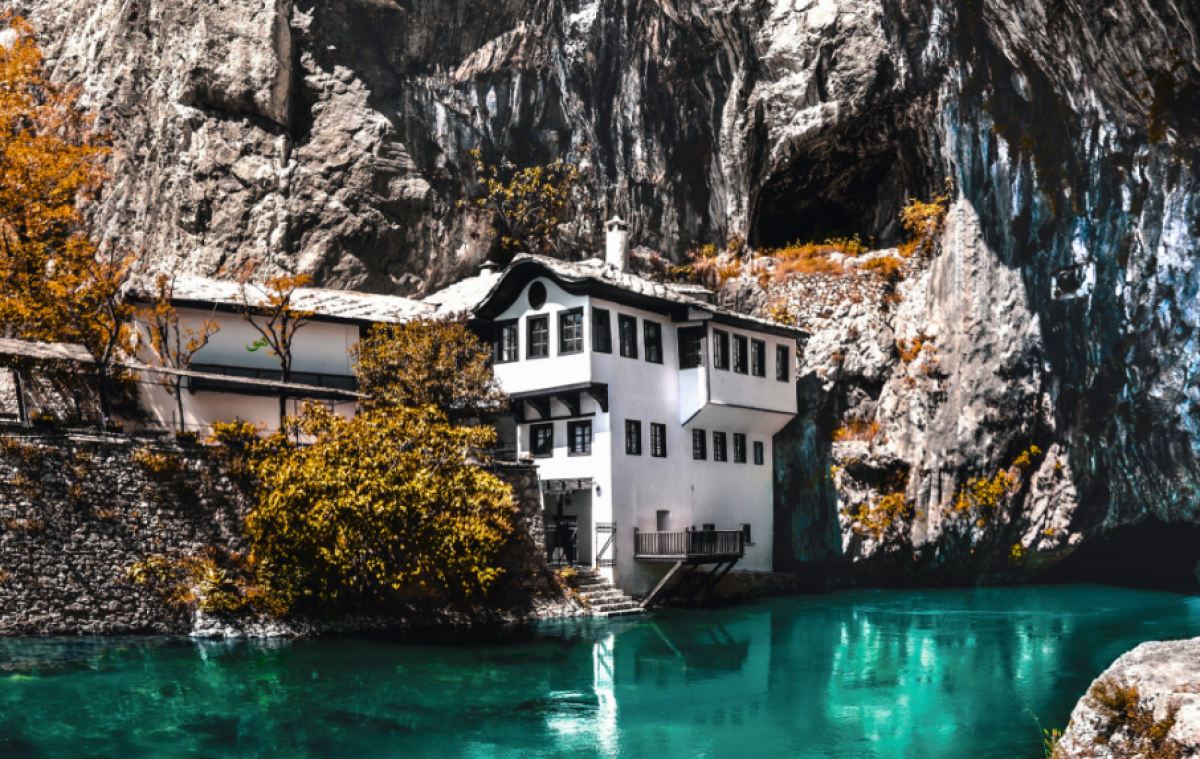 Eingefügt zwischen der hohen Felswand und dem Wasser des blau-grünen Flusses steht ein schmales, hohes Haus mit weißer Fassade und vielen dunkelbraunen Holzfenstern.