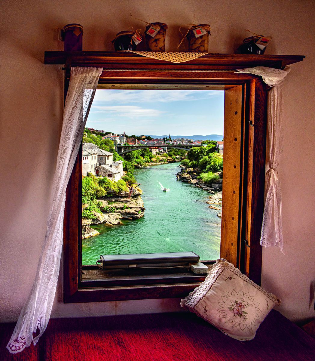 Blick aus einem Fenster auf die Stadt und den gleichnamigen Fluss Neretva. Auf dem Fluss fährt ein Motorboot, in einiger Entfernung überspannt ihn eine hohe Brücke. Im Raum sehen wir um das alte Holzfenster einen flattrigen Spitzenvorhang, ein umhäkelter 