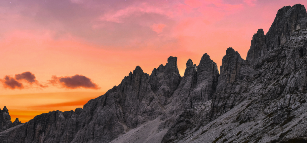 Alpenglühen über den schroffen Felszacken des Dolomitengebirges: Der Himmel verläuft von unten nach oben in Orange-, Gelb- über Lachs-, Rosa-, Pink- bis Violettönen.