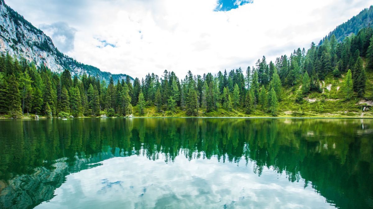 Ein See am Fuß des Gebirges, die Wälder und der Himmel spiegeln sich auf der Oberfläche des moosgrünen Gewässers.