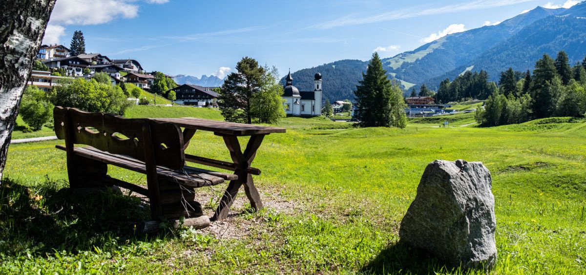 Im Vordergrund eine Holzbank mit Tisch. Der Blick geht über eine große Wiese Richtung Seefeld und das Seekirchl mit Zwiebelturm.