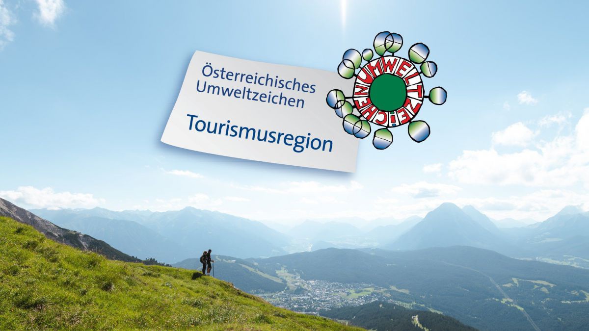 Umweltzeichen Logo eingebettet in Landschaft mit weitem Blick von einer Wiese hoch oben ins Tal. Am Bild auch 2 Wanderer.