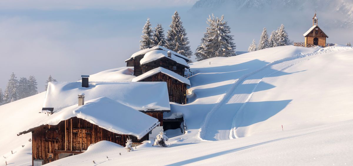 Tief verschneeite Landschaft. Holzhütten im Schnee. Eine Langlaufloipe führt vorbei. Im Hintergrund eine Kapelle aus Holz
