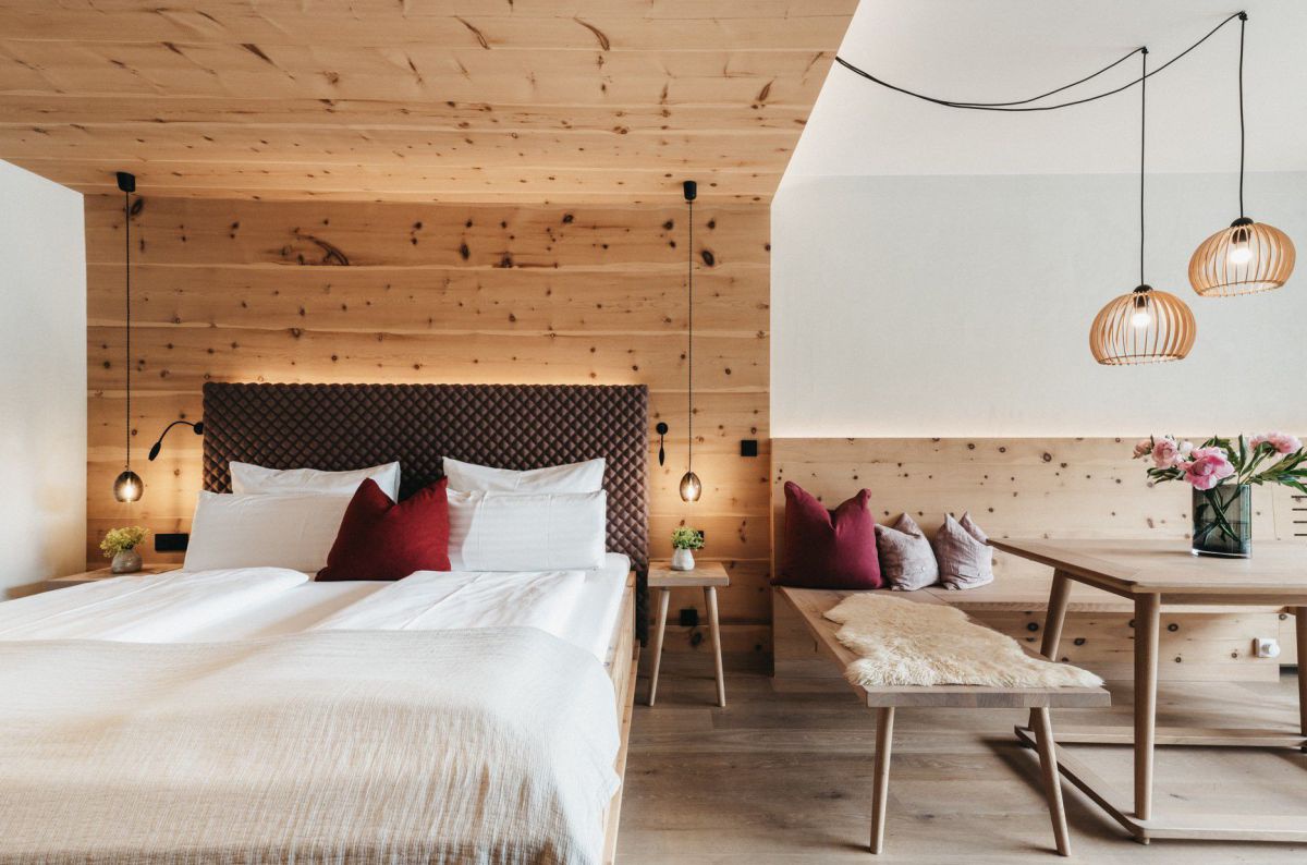 Zimmer mit Doppelbett, dahinter Holzwand, rechts ein Holztisch mit Holzbank