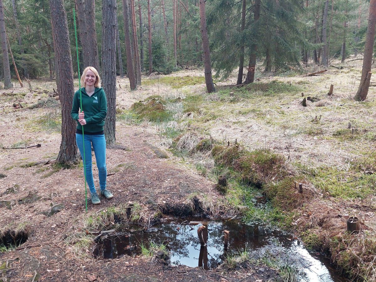 Frau Ebhart steht mit der Lawinensonde nebeen einem Tümpel im Wald.