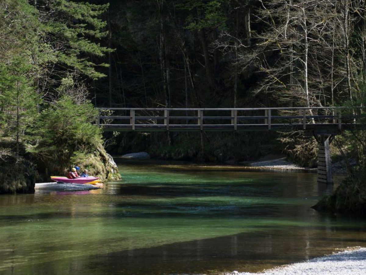 Ein bemanntes/befrautes Kajak auf dem Fluss Salza, über den sich eine Brücke spannt. Das Ufer ist bewaldet. Das Wasser schimmert smaragdgrün wo die Sonne hinfällt.
