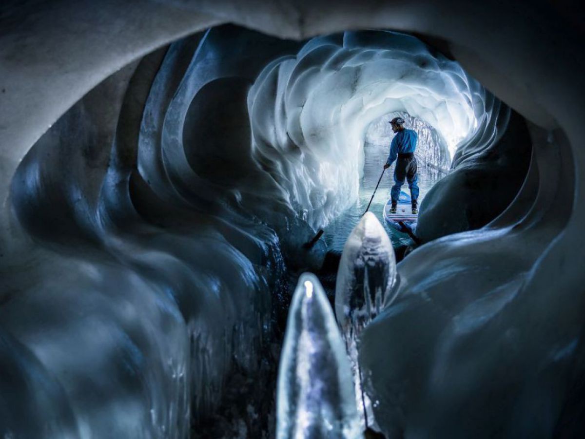 Im Inneren einer Eishöhle, durch deren Gänge sich ein Gletschersee verzweigt: Die eisigen Wände schimmern bläulich, ein Mann erkundet auf einem Stand-Up Paddle die Höhle.