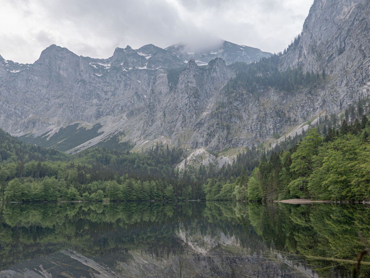 Blick auf den See inmitten der hohen Berge. Wald und Bergpanorama spieglen sich in der glatten Wasserfläche. Die Gipfel sind teilweise nebel- oder wolkenverhangen.