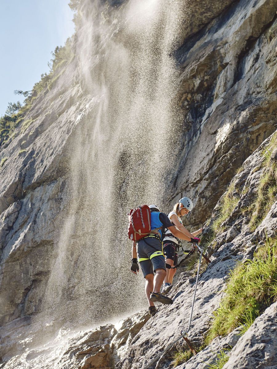 Zwei Wanderer besteigen einen felsigen Hang, über dem die Gischt eines Wasserfalls fällt. Dem Licht nach ist es früher Nachmittag.