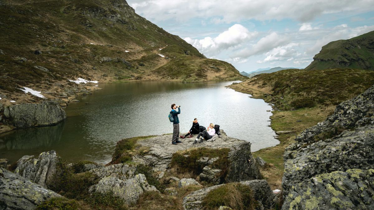 Gruppe von drei Personen bei einer Rast auf einem Felsen bei einem Gebirgssee.