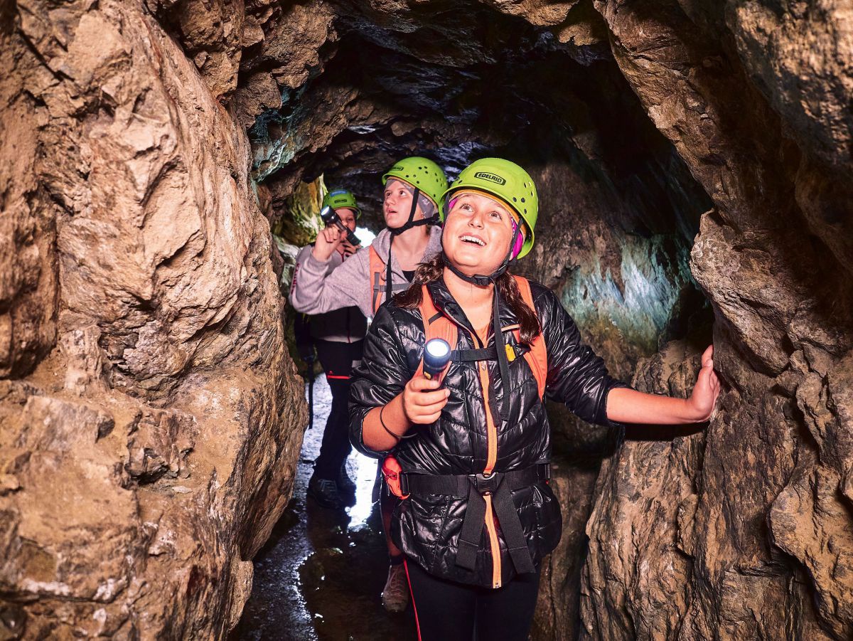 3 Personen gehen mit Taschenlampe durch eine enge, feuchte Höhle.