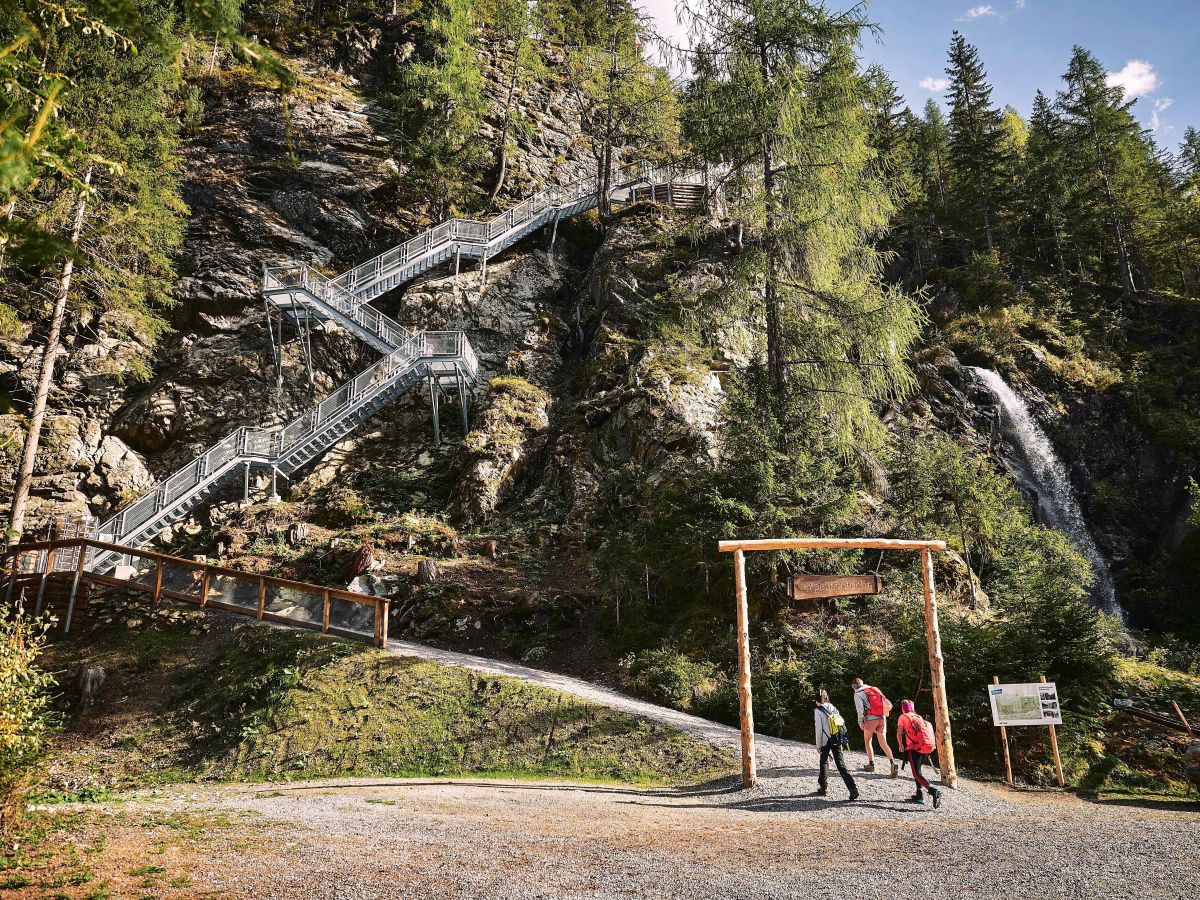 3 Wanderer gehen durch einen Holzbogen, im Hintergrund eine lange Stiege aus Stahl, rechts ein Wasserfall. 