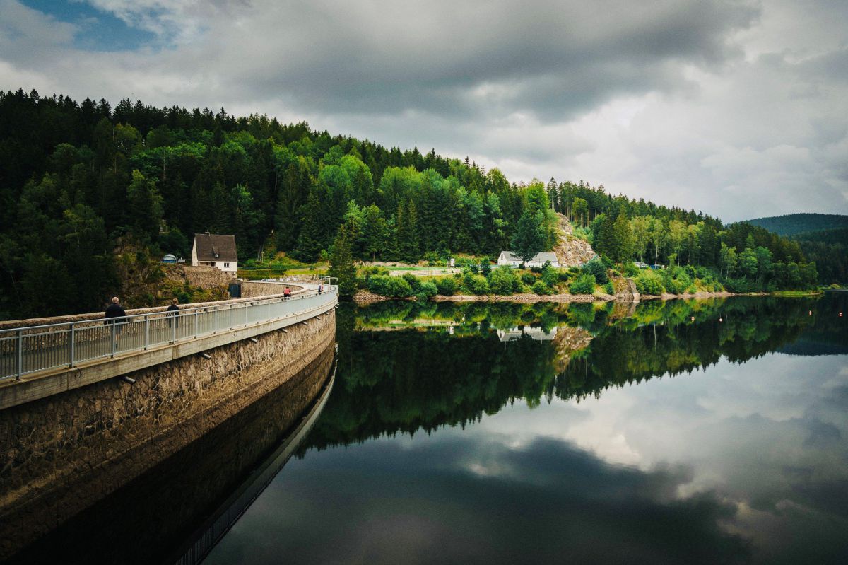 Radfahrer überqueren eine befahrbare Dammmauer, rechts im Bild spiegelt sich der wolkenverhangene Himmel im spiegelglatten Wasser des Stausees. Am Horizont ein bewaldeter Hügel, an dessen Fuß man kleine Häuschen erkennen kann.
