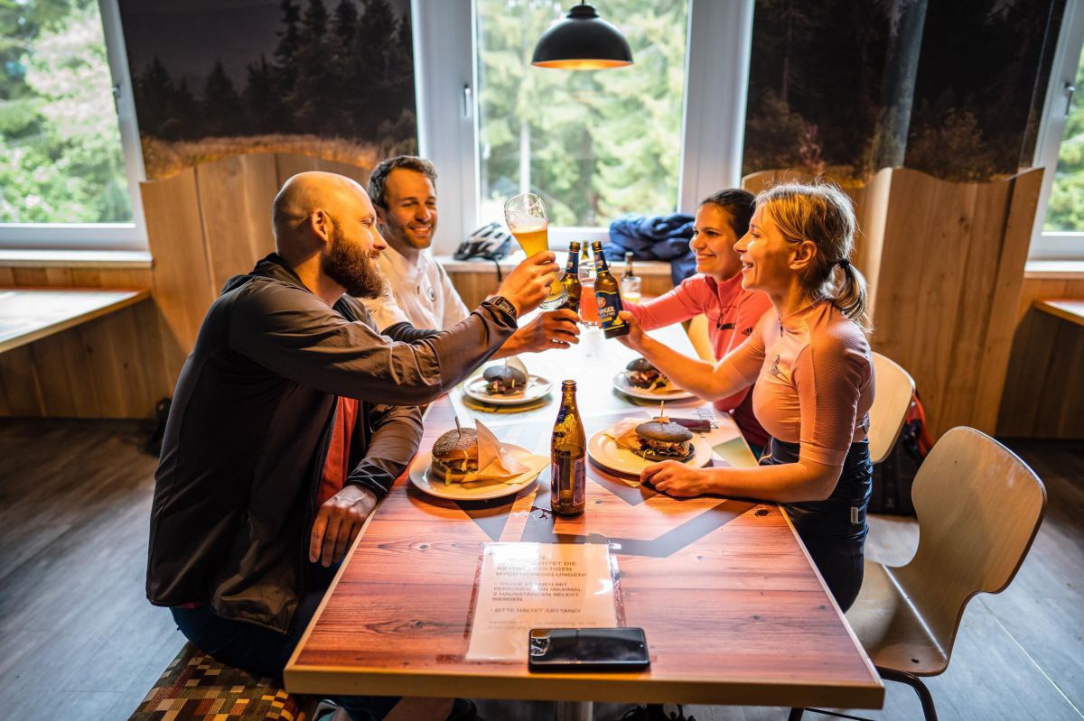 Zwei Männer und zwei Frauen sitzen einander gegenüber in einer Gastwirtschaft und stoßen mit Bier an. Auf den Tellern vor ihnen warten hausgemachte Burger darauf, verzehrt zu werden.