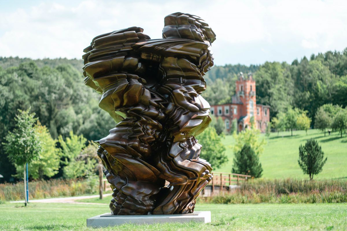 In einer naturnah gepflegten Parkanlage steht eine abstrakte Skulptur aus Bronze. Im Hintergrund ein Schloss und dahinter viel Wald.