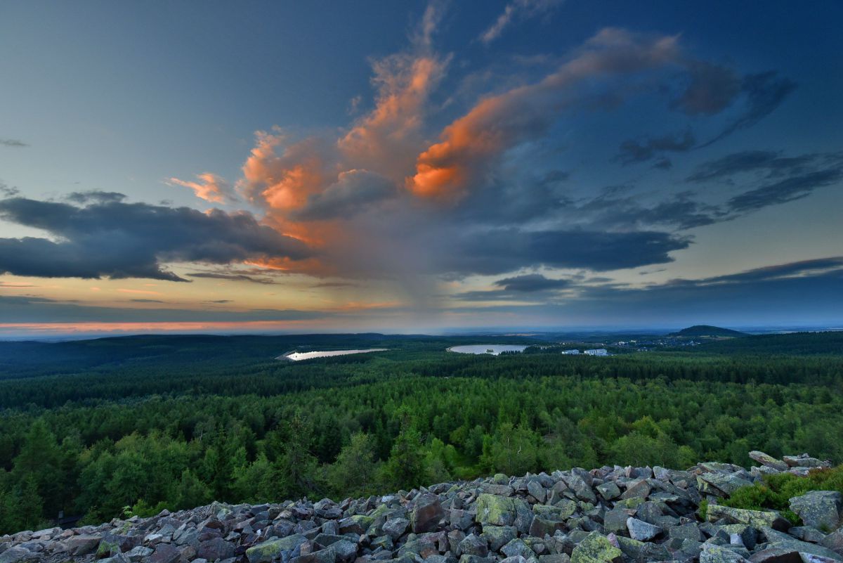 Ausblick von einem erhöhten, felsigen Punkt auf ein weites Tal aus Wäldern und Wasserflächen in einiger Entfernung. Der Himmel ist von dunkeln Wolken durchzogen, welche teilweise durch das Licht der untergehenden Sonne rotgolden leuchten.