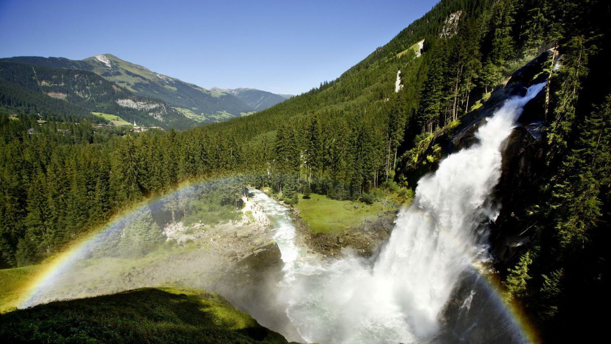 Ein Wasserfall in bewaldeter Berglandschaft, im Sprühnebel des Wasserfalls ist ein Regenbogen sichtbar.