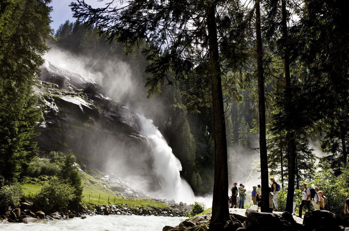 Blick aus dem Wald auf den Wasserfall. Neben dem Fluss hält sich eine Gruppe Besucher*innen auf, die den Blick auf den Wasserfall und die wohltuende Gischt genießen.
