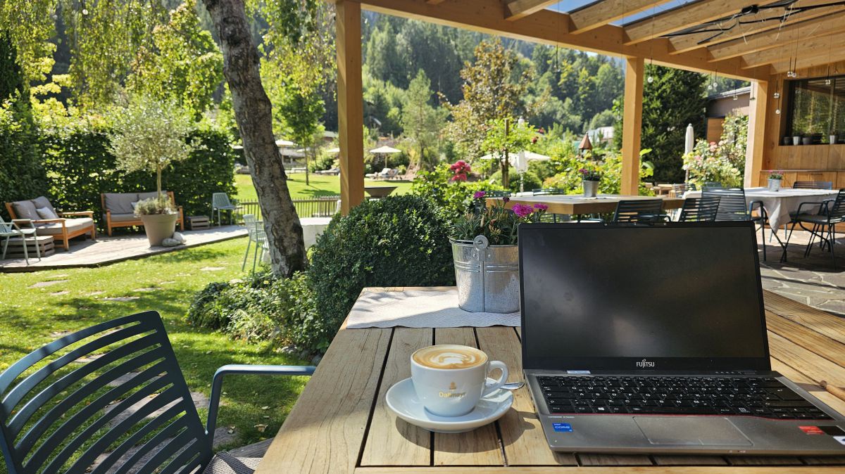 Laptop und Kaffee auf der Terasse, im Hintergrund ein Garten mit üppigem Grün.