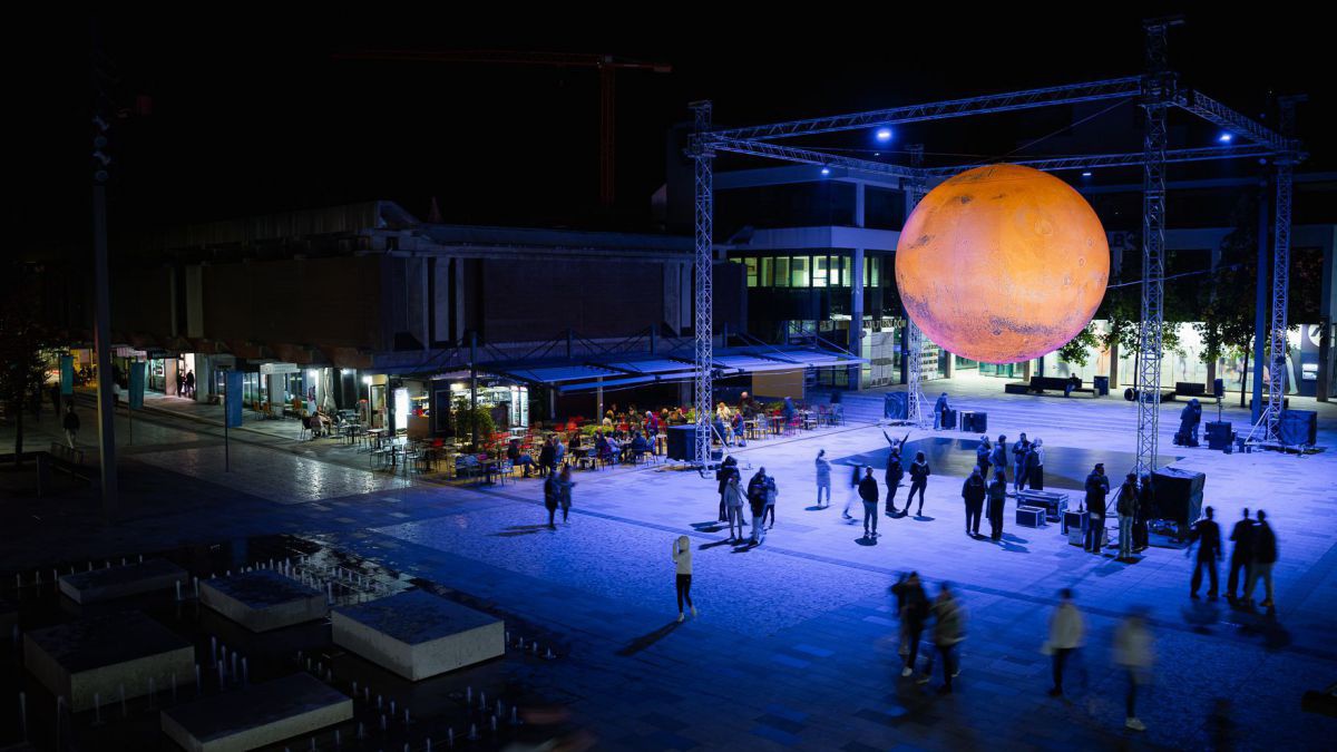 Es ist Nacht. Zwischen einem Stahlgerüst schwebt eine große orange Kugel.