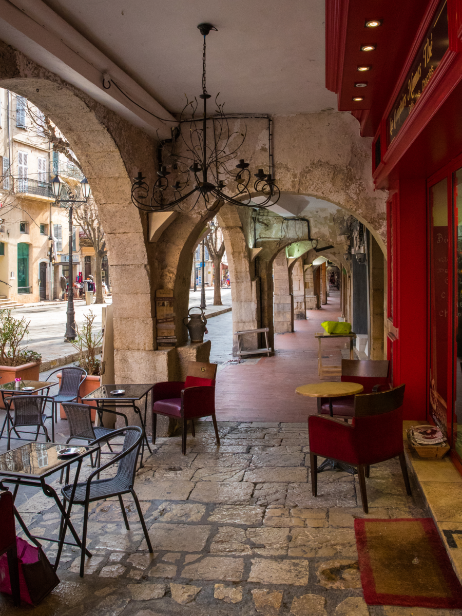 Ein Gastbereich mit Gruppen von Tischen und Stühlen in einem historischen Arcadengang. Die Fassade des Restaurants ist aus rotem Holz, von der Decke hängt ein alter schmiedeeisender Luster mit leeren Kerzenhaltern.