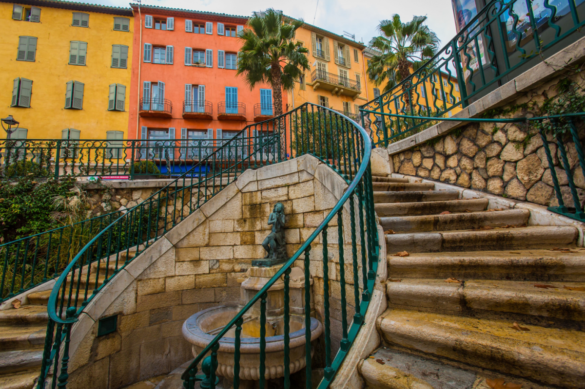 Blick vom Fuße einer geschwungenen Doppeltreppe mit Brunnenund grünen Eisengeländern hinauf auf Hausfassaden in kräftigen Gelb- und Orangetönen mit blauen Holzfensterläden.