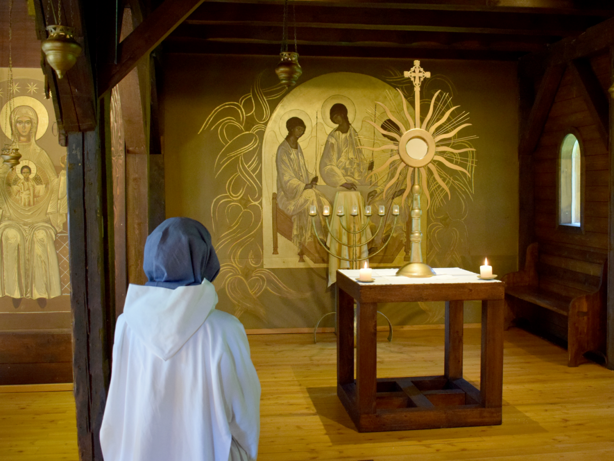  Im Inneren einer Kapelle steht eine Klosterfrau im Habit vor einem schlichten Altar mit Monstranz, dahinter ein Goldenes Altarbild.