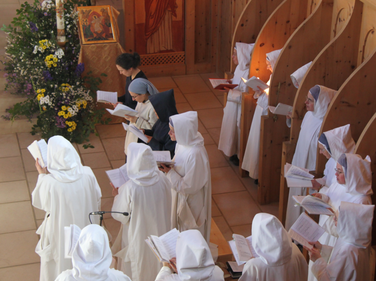Eine Gruppe Klosterschwestern in weißen Habits stehen im Halbkreis in der Kapelle und halten Gebetsbücher in den Händen, woraus sie etwas lesen oder singen.