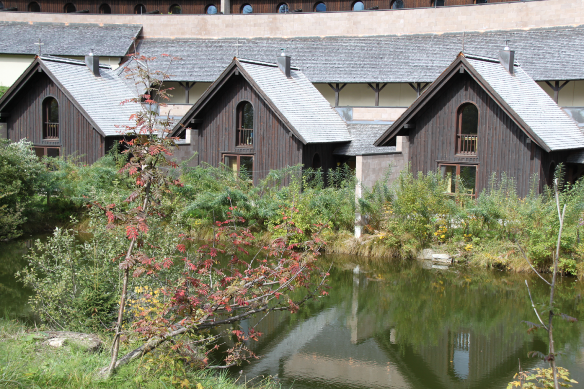 Mehrere sehr kleine Holzhütten mit kleinen Vorgärten am Ufer eines Gewässers. Im Hintergrund sieht man das Hauptgebäude des Klosters.
