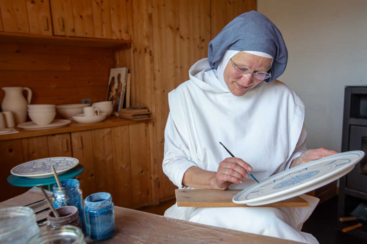 Eine der Klosterfrauen beim Bemalen eines Keramiktellers. Hinter ihr auf einer Anrichte aus Nadelholz stehen mehrere noch rohe, unbemalte Gefäße. Neben ihr auf dem Tisch halb fertige Stücke und leere Marmeladegläser voll Glasur mit Pinseln darin.