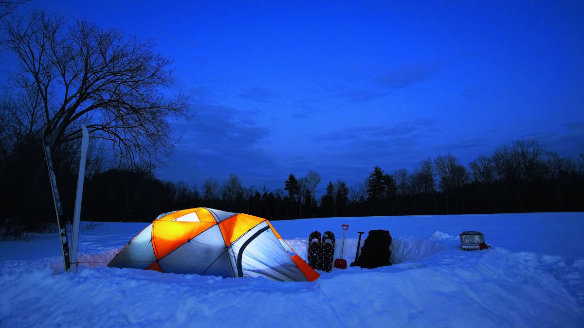 Ein von innen beleuchtetes Zelt auf einer verschneiten Wiese am Waldrand. Der Himmel ist dunkelblau, das Zelt leuchtet hellblau und orange. Neben dem Zelt stehen Schneeschuhe, Rucksack, Schneeschaufeln.