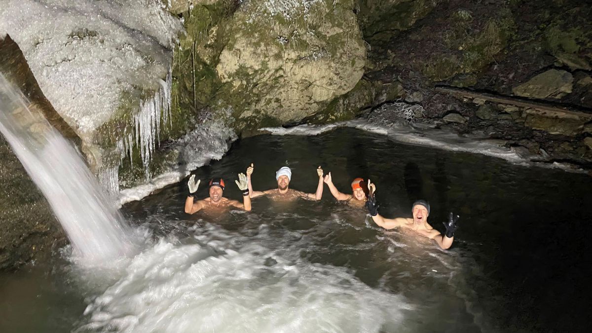 4 Personen in einem Gewässer bei einem Wasserfall. Es ist finster. Am Hang hängen Eiszapfen herunter, am Ufer liegt Schnee