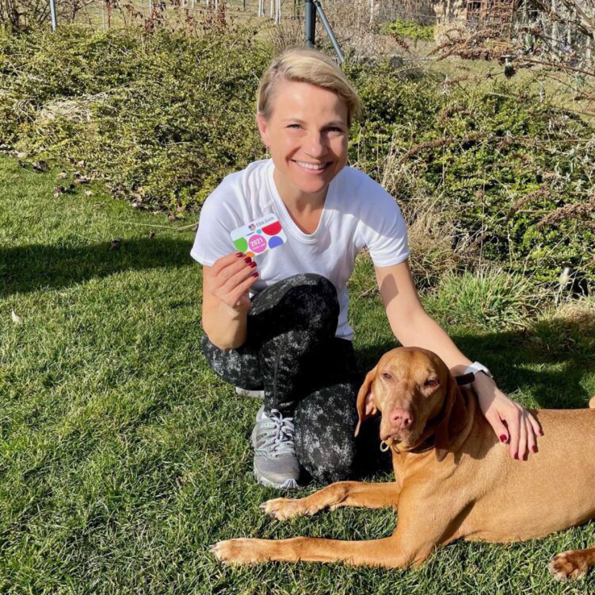 Kristina Sprenger hockt auf einer Wiese, streichelt einen Hund und hält die Ethik Card in der Hand.