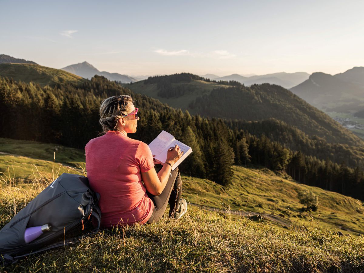 Frau sitzt mit einem Buch auf einer Bergwiese in der Abendsonne.