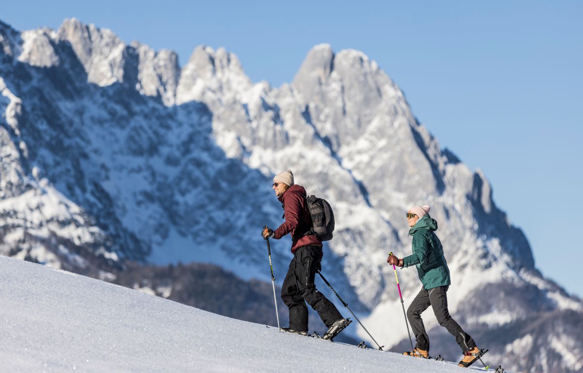 Mann und Frau steigen einen schneebedeckten Hang hoch, im Hintergrund der Wilde Kaiser.