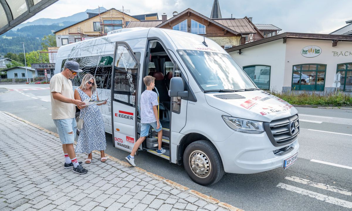 Bub steigt in einen Kleinbus, davor studiert ein Paar eine Wanderkarte.