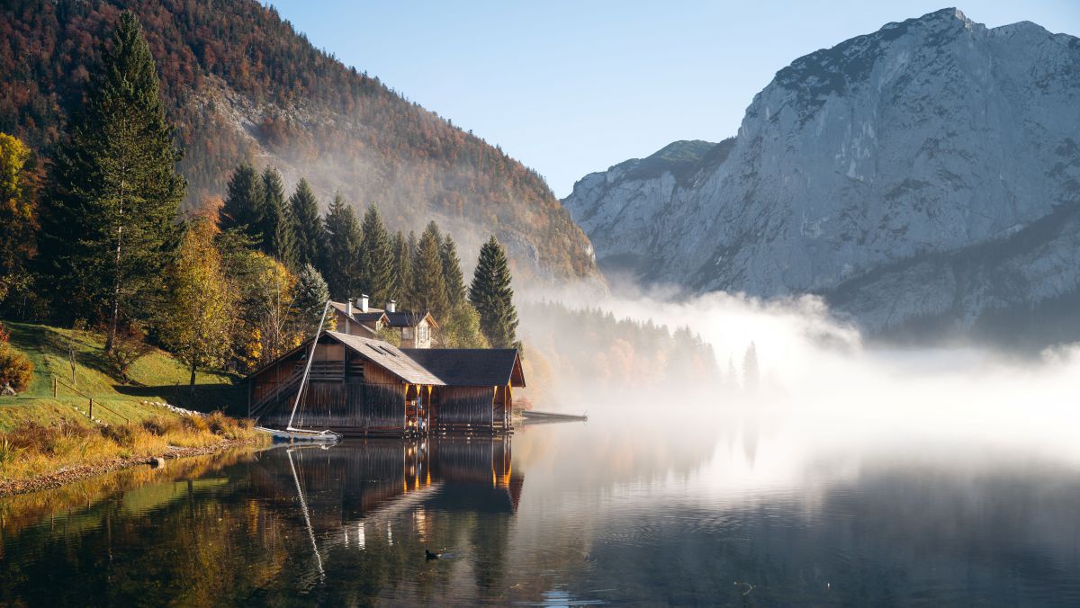 See, am Ufer ein Haus, Bäume sind herbstlich gefärbt, am Wasser liegt Nebel.