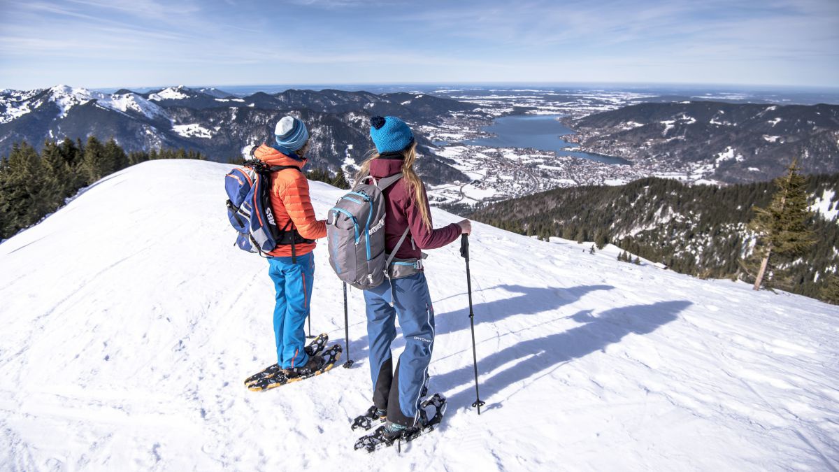2 Personen auf Schneeschuhen blicken von einer Anhöhe hinunter auf einen See.