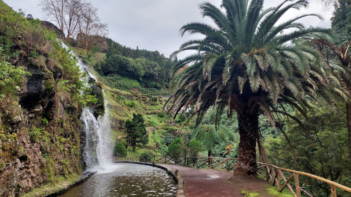 Wasserfall, das Wasser fällt in einen gemauerten Teich, daneben ein Weg und eine  Palme.