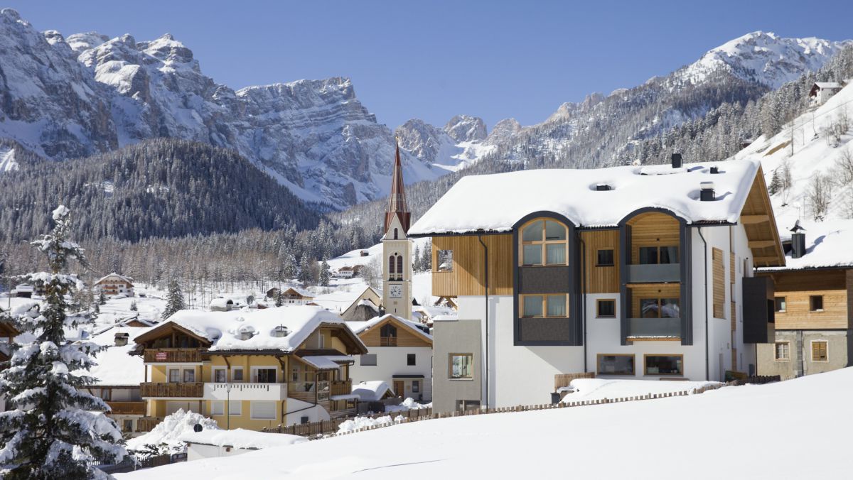 Blick auf das Hotel im tief verschneiten Winter, dahinter der Ort mit dem spitzen Kirchturm und dahinter die mächtige Bergkulisse der Dolomiten.