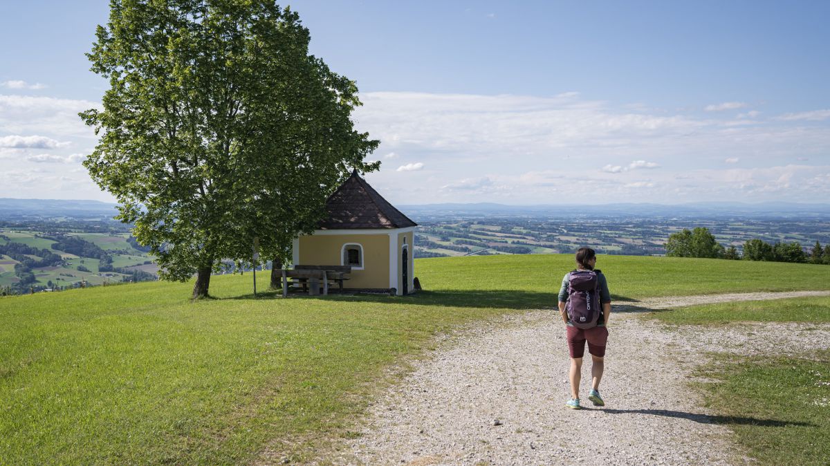 Frau wandert auf einem breiten Weg vorbei an einer Kapelle im Schatten eines Baumes.
