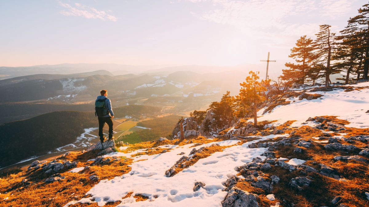 Mann in der Abendsonne auf einer Anhöhe mit weitem Blick ins Tal, am Boden noch einige Schneefelder.