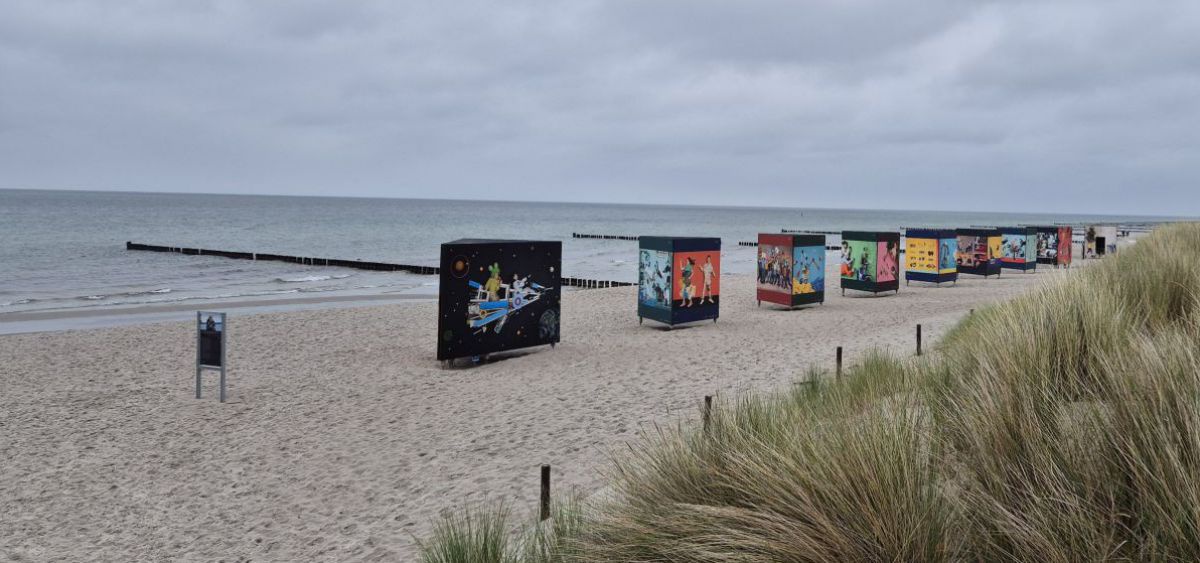 Ein Sandstrand unter tief bewölktem Himmel, ein Teil ist hoch mit Riedgras bewachsen. Den Strand entlang ist eine Kunstausstelllung mit Bildern aufgebaut.