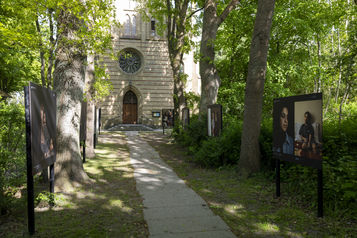 Fotowände säumen abwechselnd mit grün belaubten Bäumen einen Weg, an dessen Ende die Kirche mit dem Eingangstor und darüberliegendem Rosettenfenster im neugotischen Stil steht.
