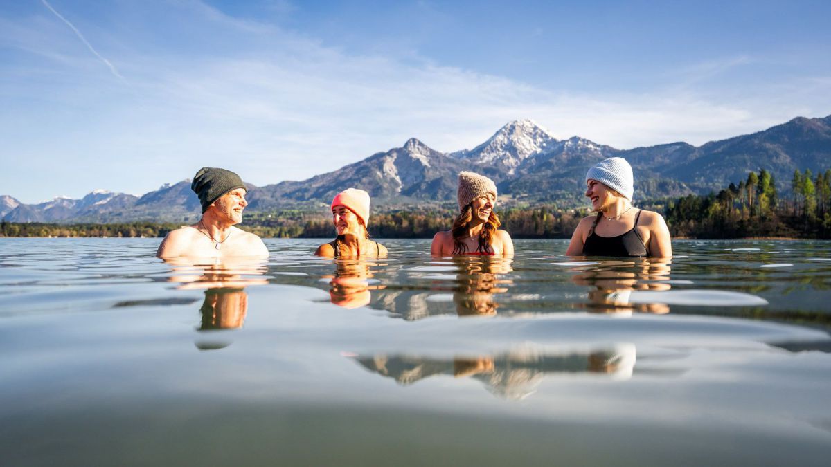 4 Personen stehen im Wasser, alle haben eine Haube auf, hinter dem See eine Bergkulisse.