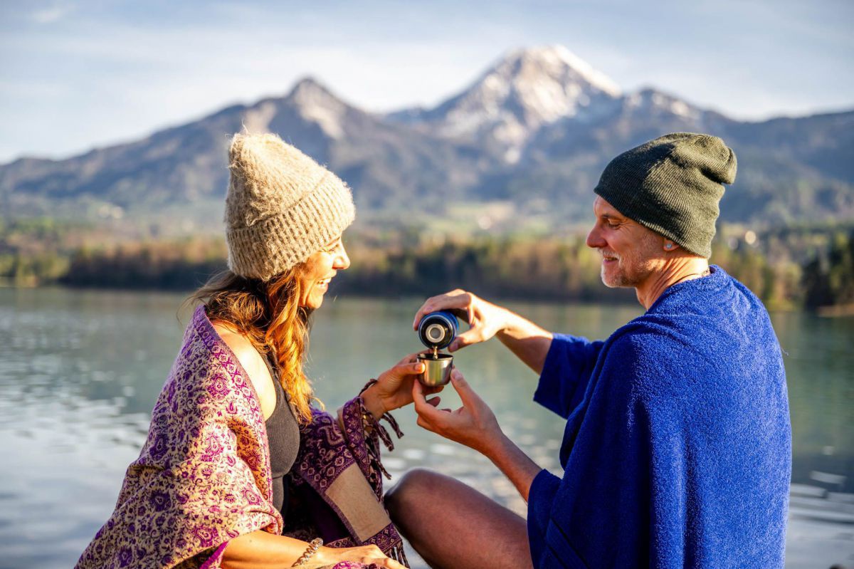 Mann und Frau sitzen in einem Handtuch gehüllt am Ufer. Mann schenkt der Frau Tee ein.